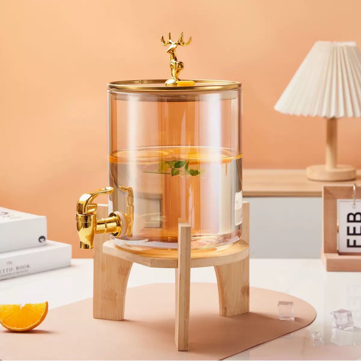Clear glass drink dispenser with gold accents on a wooden stand against a peach-colored wall.